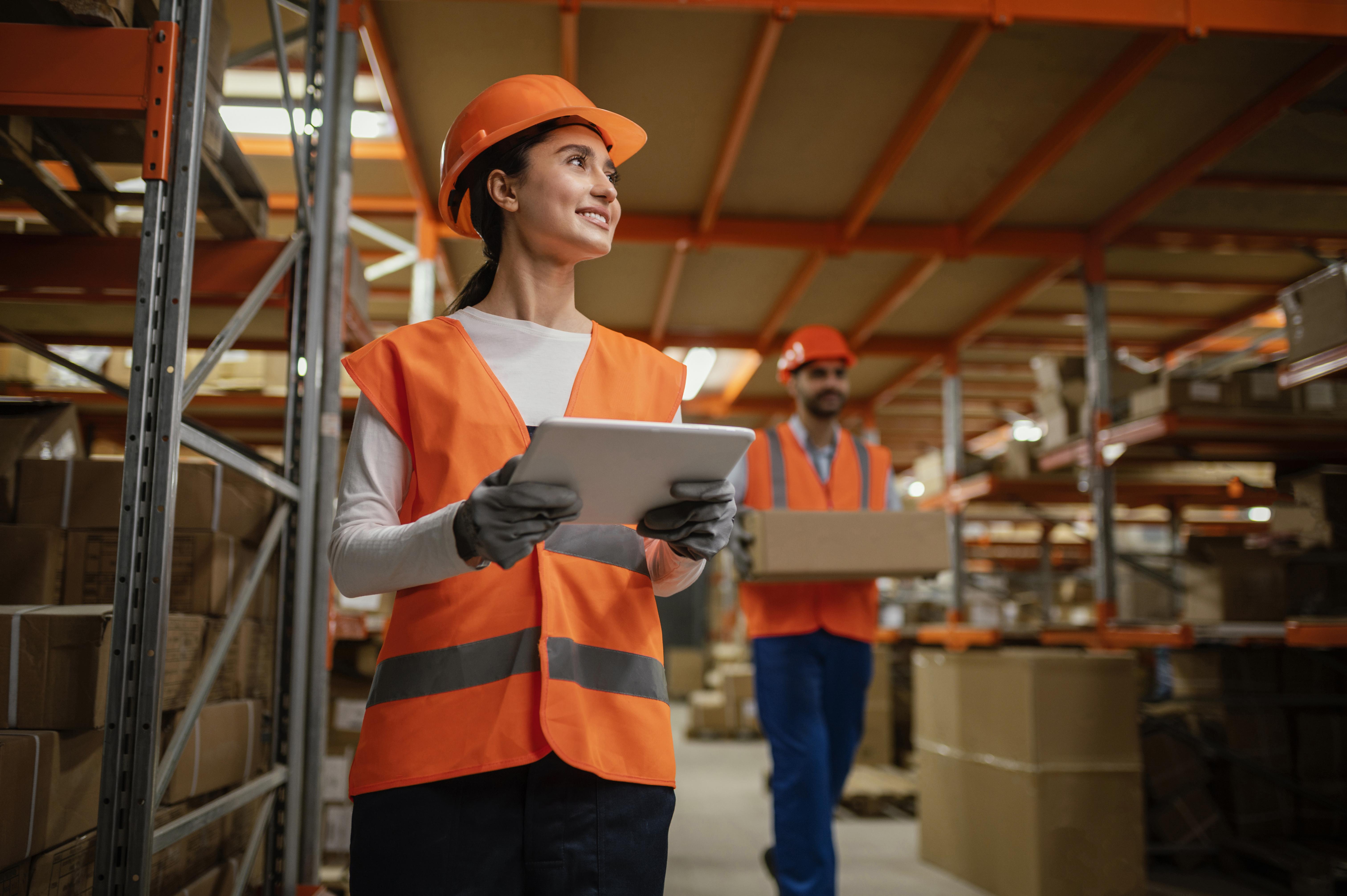 Professional woman in safety vest and hard hat using tablet in modern warehouse environment